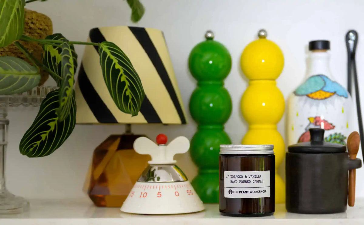 A styled shelf display featuring a 'Tobacco & Vanilla' hand poured candle by The Plant Workshop in a dark amber glass jar, positioned next to a black ceramic pot with a wooden spoon, a white bottle with a colorful illustration, and a small white and red bird-shaped object.