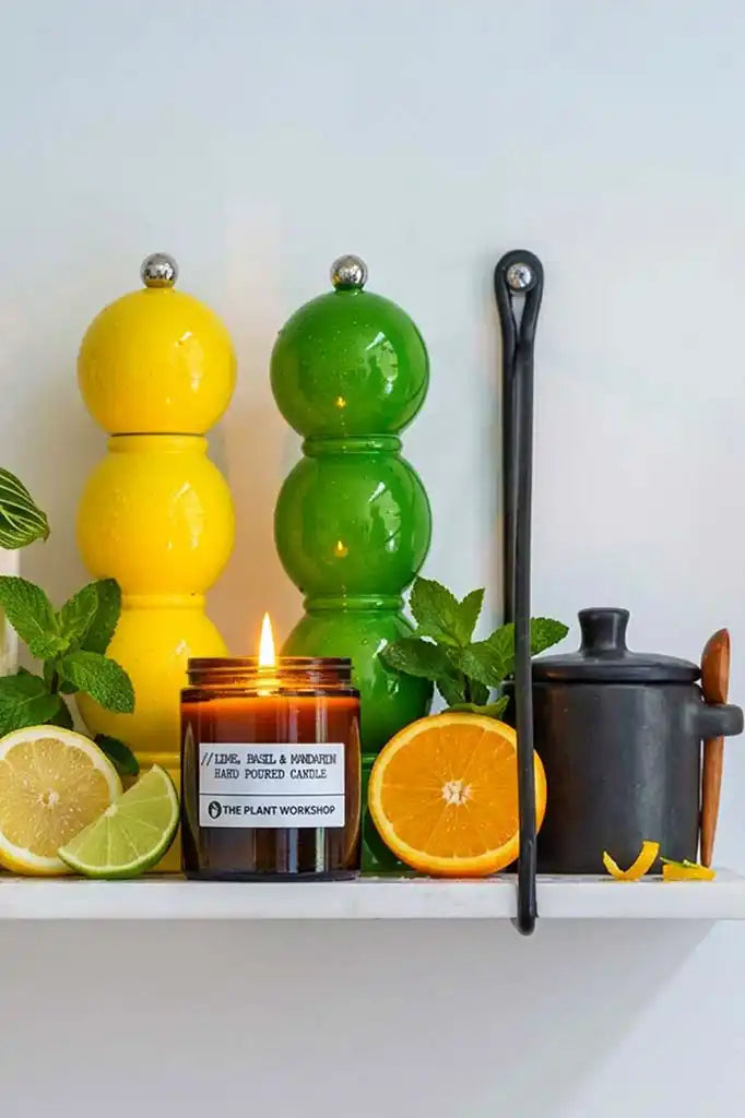 A lit amber glass candle jar labeled 'The Plant Workshop' sits on a white shelf, surrounded by citrus slices, mint leaves, two glossy ceramic pepper mills in yellow and green, a black spoon, and a black lidded pot against a plain light wall.