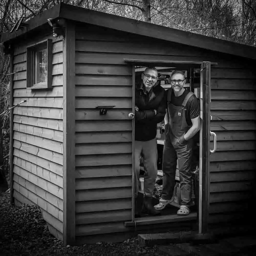 A black and white photograph of two men standing in the open doorway of a wooden shed. The shed has horizontal siding and a small window on the side.