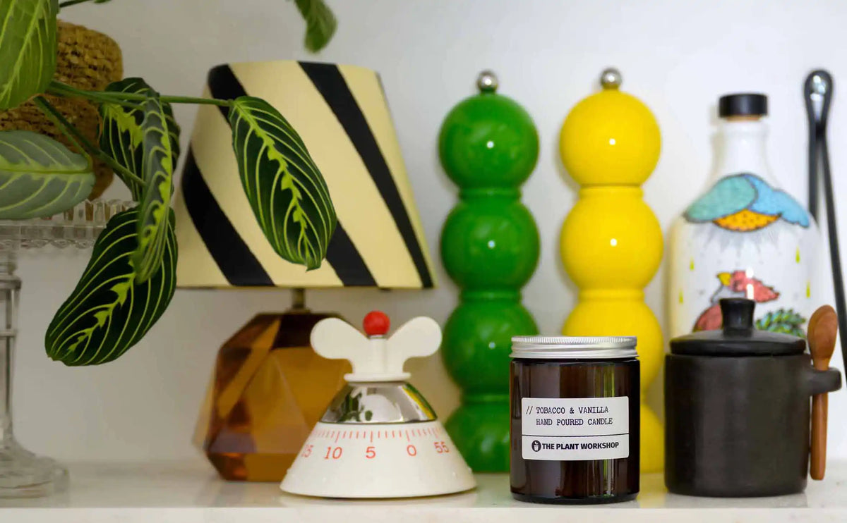 A styled shelf display featuring a 'Tobacco & Vanilla' hand poured candle by The Plant Workshop in a dark amber glass jar, positioned next to a black ceramic pot with a wooden spoon, a white bottle with a colorful illustration, and a small white and red kitchen timer.