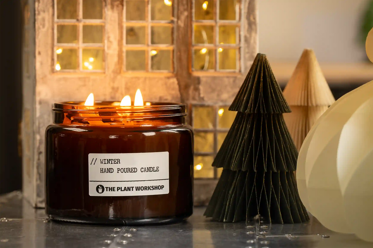 A lit amber glass jar candle with a white label reading 'WINTER HAND POURED CANDLE' and 'THE PLANT WORKSHOP' sits on a tabletop in a styled setting with warm fairy lights, a wooden window backdrop, paper tree decorations, and a cream paper lantern.